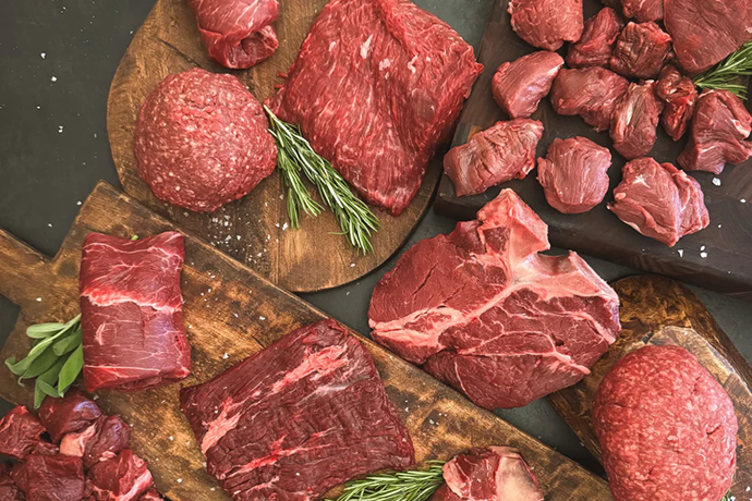 Raw beef cuts and chunks arranged on wooden cutting boards with rosemary sprigs, photographed from above on a dark surface.